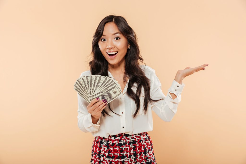 Excited Young Lady With Long Dark Hair Holding Fan 100 Dollar Bills Expressing Gladness Having Lot Money Peach Background